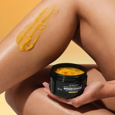 Person applying The Body Shop's Refreshing Passionfruit gel body scrub to their leg with a jar of the product in their hand